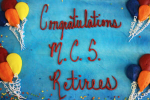 Blue cake with "Congratulations MCS Retires" written in red icing. There are yellow, orange, blue and red balloons, made of icing on the sides of the cake.